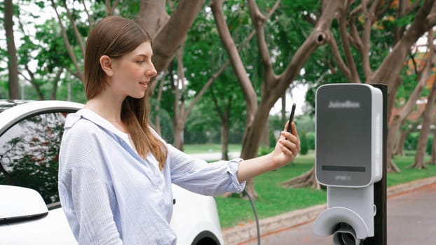 Woman using smartphone online banking application to pay for electric car battery charging from EV charging station during vacation holiday road trip at national park or summer forest. Exalt