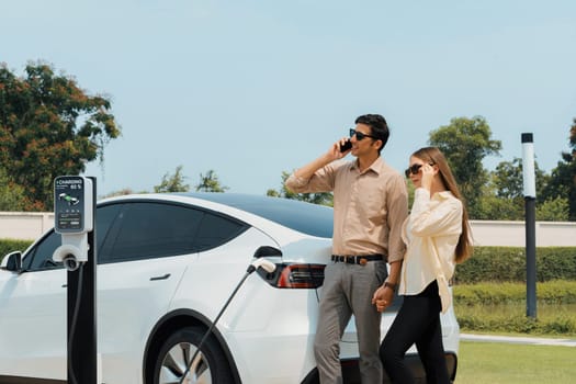 Young couple recharge her EV electric vehicle at green city park parking lot while talking on phone. Sustainable urban lifestyle for environment friendly EV car with battery charging station.Expedient