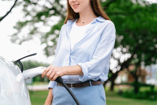 Young woman recharge EV electric vehicle's battery from EV charging station in outdoor green city park scenic. Eco friendly urban transport and commute with eco friendly EV car travel. Exalt