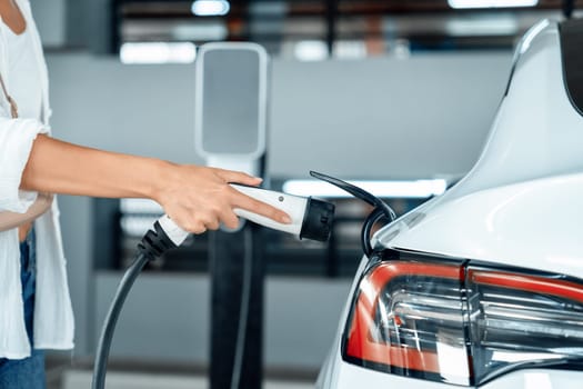 Young woman travel with EV electric car to shopping center parking lot charging in downtown city showing urban sustainability lifestyle by green clean rechargeable energy of electric vehicle innards