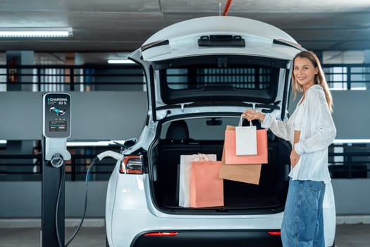 Young woman travel with EV electric car to shopping center parking lot charging in downtown city showing urban sustainability lifestyle by green clean rechargeable energy of electric vehicle innards