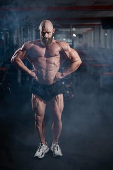 Muscular bald man posing in shorts. Bodybuilder showing off his shape in the gym