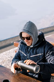 Man in a jacket with a pulled hood pours coffee from a thermos into a mug while sitting at a table in the mountains. High quality photo
