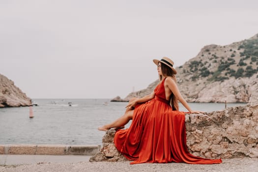 Side view a Young beautiful sensual woman in a red long dress posing on a volcanic rock high above the sea during sunset. Girl on the nature on overcast sky background. Fashion photo