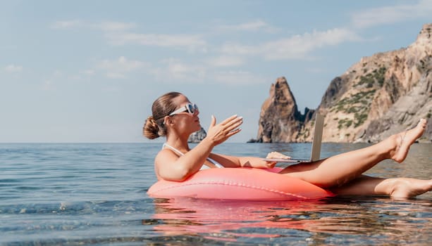 Woman freelancer works on laptop swimming in sea on pink inflatable ring. Pretty lady typing on computer while floating in the sea on inflatable donut at sunset. Freelance, remote work on vacation