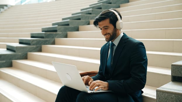 Professional business man sitting at stairs while working on laptop. Skilled project manager listening music from headphone and checking email and discussion about marketing plan. Outdoor. Exultant.