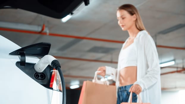 Young woman travel with EV electric car to shopping center parking lot charging in downtown city showing urban sustainability lifestyle by green clean rechargeable energy of electric vehicle innards