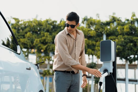 Young man recharge electric car's battery from charging station in outdoor green city park in springtime. Rechargeable EV car for sustainable environmental friendly urban travel lifestyle. Expedient