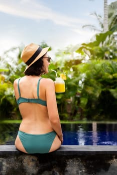 Back view of young woman on vacation drinking pineapple juice relaxing sitting on swimming pool ledge in tropical holiday resort. Vertical image. Summer vacation concept.
