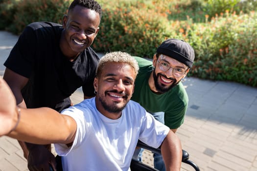 Young black man in a wheelchair using phone taking selfie together with male friends outdoors. Friendship and technology concept.
