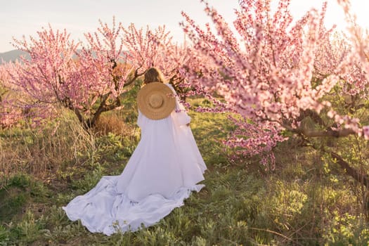 Woman blooming peach orchard. Against the backdrop of a picturesque peach orchard, a woman in a long white dress and hat enjoys a peaceful walk in the park, surrounded by the beauty of nature