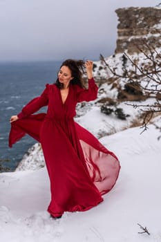 Woman red dress snow sea. Happy woman in a red dress in the snowy mountains by the emerald sea. The wind blows her clothes, posing against sea and snow background