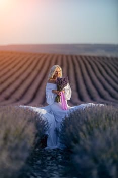 Blonde woman poses in lavender field at sunset. Happy woman in white dress holds lavender bouquet. Aromatherapy concept, lavender oil, photo session in lavender.