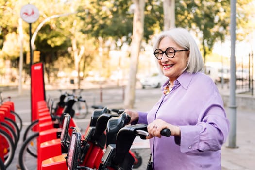 smiling senior woman taking rental bike from parking row, concept of sustainable mobility and active lifestyle in elderly people