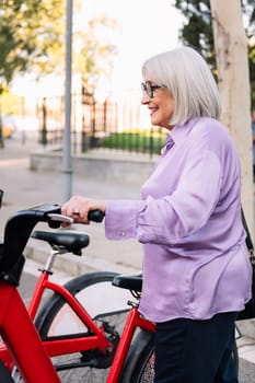 smiling senior woman taking rental bike from parking row, concept of sustainable mobility and active lifestyle in elderly people