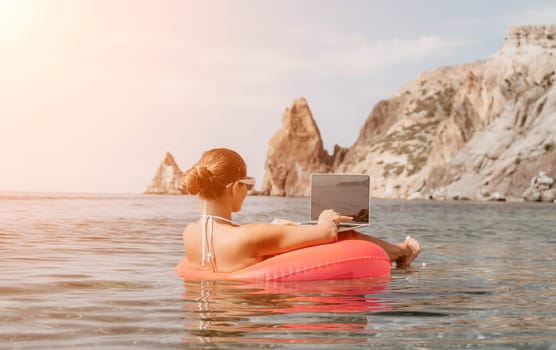 Woman freelancer works on laptop swimming in sea on pink inflatable ring. Pretty lady typing on computer while floating in the sea on inflatable donut at sunset. Freelance, remote work on vacation