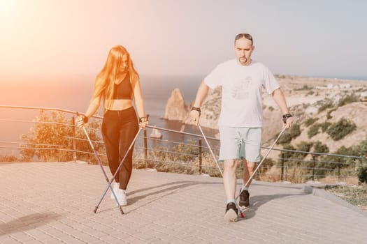 Happy Middle aged couple or friends practicing nordic walking in park near sea. Mature couple with trekking poles walking, practicing Nordic walking outdoors. Aging youthfully and sport concept.