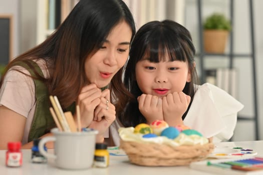 Happy mother and little daughter having fun coloring and decorating eggs for Easter holiday at home.