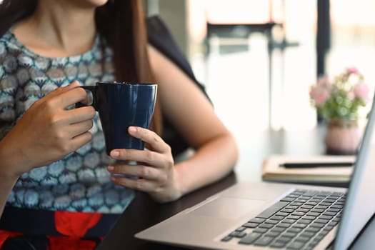 Businesswoman holding cup of coffee and reading email on her laptop screen.