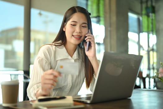 Pleased young businesswoman holding credit card and talking on mobile phone with bank customer service.