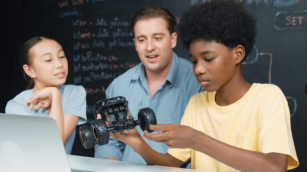 Smart teacher inspect car model construction to diverse student. Highschool children with mixed races listening mentor explain robotic model system at table with laptop and wires placed. Edification.