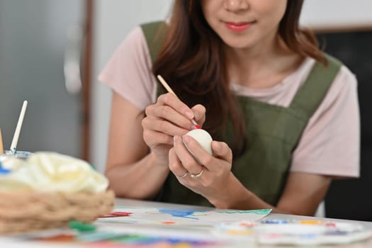 Smiling young woman in apron painting Easter egg with brush at table. Preparing for Easter concept.