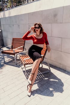 Portrait of a woman on the street. An attractive woman in glasses, a red blouse and a black skirt is sitting on a bench outside