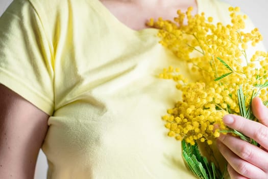 Close up young girl or woman in yellow dress holds yellow brunch of mimosa flowers. 8 march women's day concept.