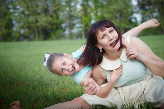 Happy mother and daughter enjoying rest, playing and fun on nature on a green lawn and with blooming apple tree in the background. Woman and girl resting outdoors in summer and spring day