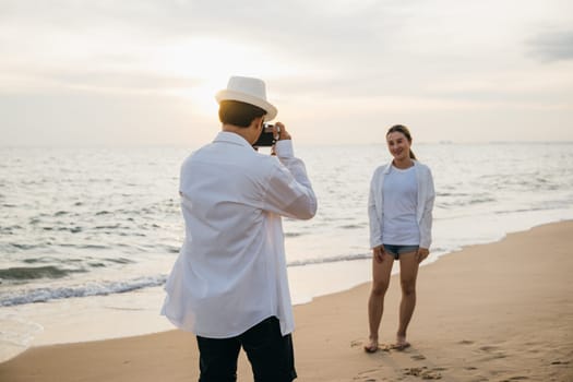 Romantic sunrise at beach for young couple in white as boyfriend takes candid photos of his girlfriend. blissful moment of togetherness filled with love and joy against backdrop of ocean.
