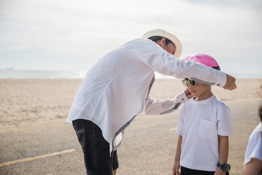 A family on vacation at the beach where the father sporting a safety helmet teaches his cheerful son the balance and joy of bicycle riding a memorable tourism day filled with family happiness.