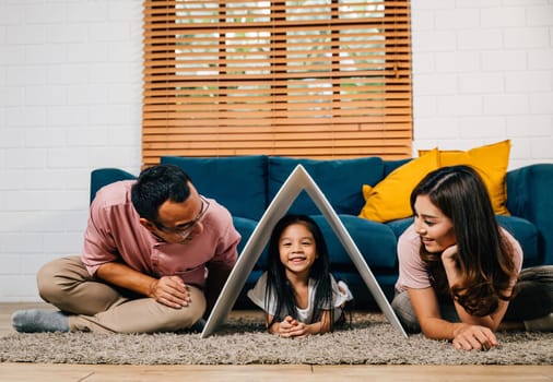 A portrait of a joyful family on a couch holding a cardboard roof in their new house symbolizing safety and support. Asian parents and their daughter exude happiness.
