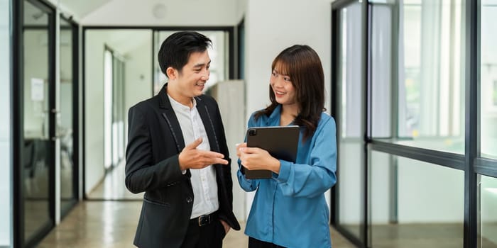 Asian woman and business man working using digital tablet tech discussing financial market data standing at corporate office.