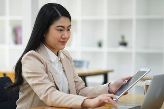 A focused businesswoman working on a laptop while holding a notebook in a bright office environment..