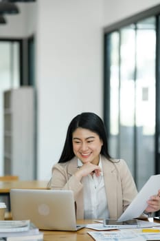 A focused businesswoman working on a laptop while holding a notebook in a bright office environment..