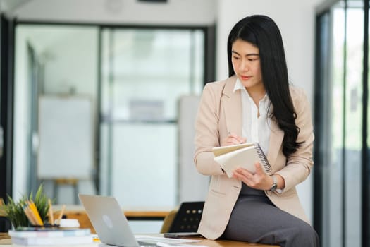 A focused businesswoman working on a laptop while holding a notebook in a bright office environment..