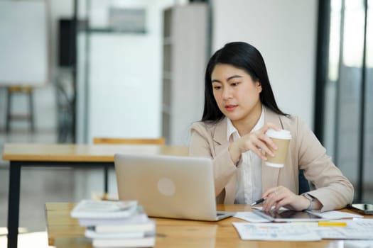 A focused businesswoman working on a laptop while holding a notebook in a bright office environment..