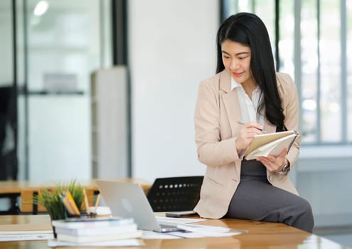 A focused businesswoman working on a laptop while holding a notebook in a bright office environment..