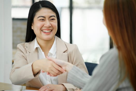 Two smiling businesswomen shaking hands over a desk in the office, symbolizing a successful agreement or partnership.