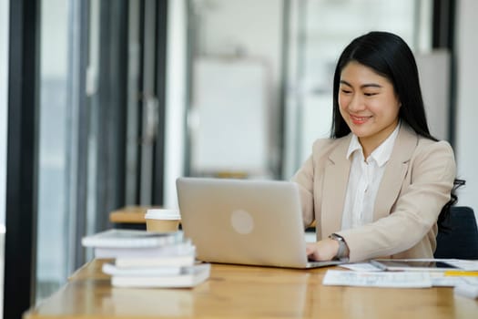 A focused businesswoman working on a laptop while holding a notebook in a bright office environment..