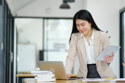 A focused businesswoman working on a laptop while holding a notebook in a bright office environment..