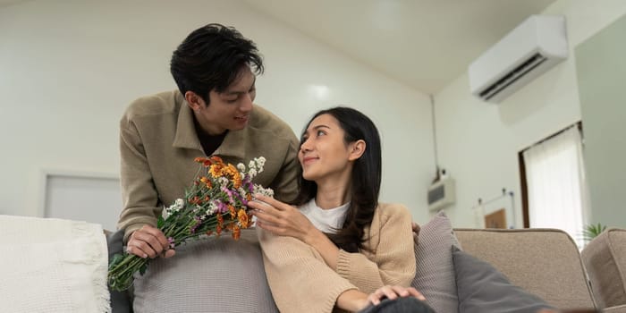 Romantic young asian couple embracing with holding flowers and smiling in living room at home. fall in love. Valentine concept.