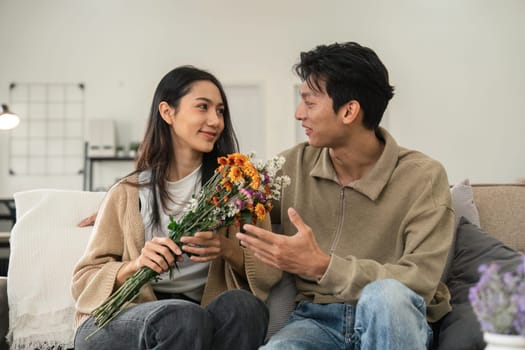 Romantic young asian couple embracing with holding flowers and smiling in living room at home. fall in love. Valentine concept.