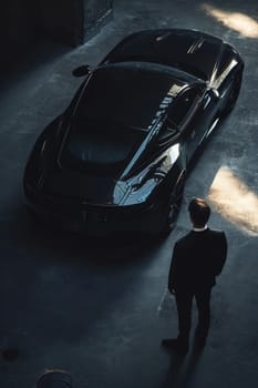 Focused man by sports car in moody lighting. A sharply dressed man in a black suit is seen from above, looking intently at sleek sports car with distinctive red rear lights in a dimly lit environment.