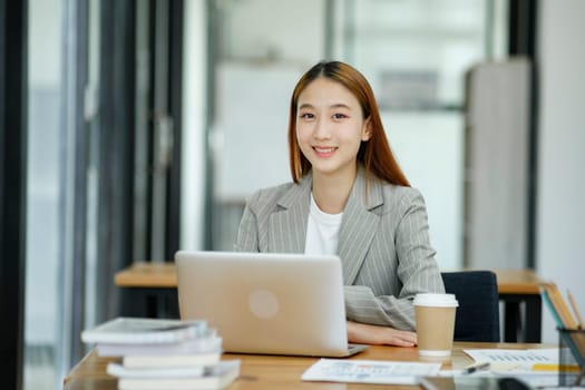 A professional and confident businesswoman smiling at the camera, standing by her desk with a laptop in a modern office.