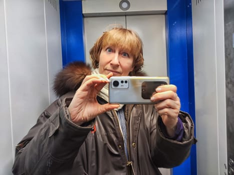 Middle-aged woman in warm jacket and a hood with fur taking selfie in mirror in elevator. Female Traveler in Alaska, Lapland, Arctic, Siberia