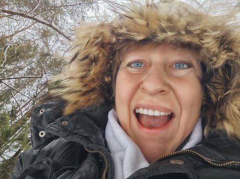 Happy, cheerful middle-aged woman in a warm jacket and a hood with fur taking selfie in winter outdoors. Female Traveler in Alaska, Lapland, Arctic, Siberia