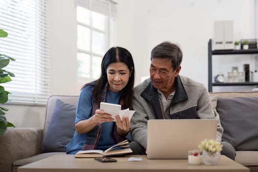 Asian senior couple checking and calculate financial bill together on sofa involved in financial paperwork, paying tax online using laptop at living room home.