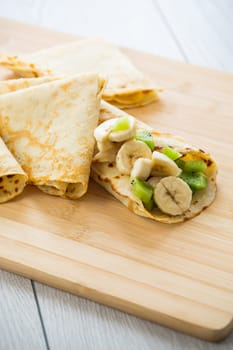 cooked thin pancakes with bananas and kiwi on a light wooden table