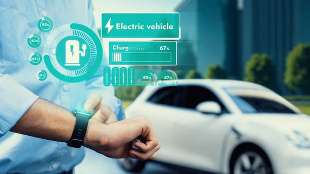 Businessman check EV car battery status on smartwatch hologram recharge from charging station ESG green park. Modern city lifestyle and eco-friendly electric car utilization to reduce CO2. Peruse